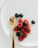 Overhead view of a white plate with a fine gold rim holding a berry cheesecake slice and a gold spoon
