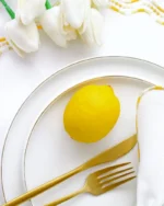White porcelain dinner plate with a fine gold rim, styled with a lemon and gold cutlery on white linen