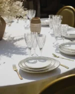 Stacked white dinner plates with gold edging on a round table with crystal glasses and gold cutlery