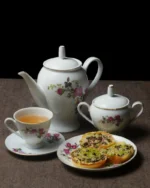 Camellia porcelain teapot, sugar bowl and teacup with saucer beside a plate of snacks
