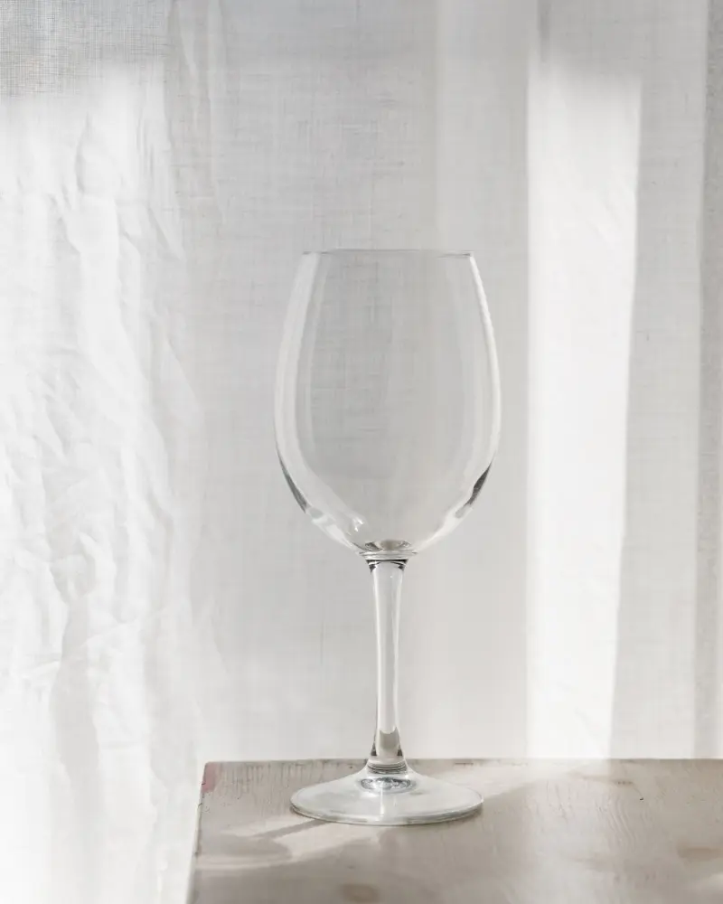 Lumière crystal wine glass on a sunlit table Empty Lumière lead-free crystal wine glass on a sunlit wooden table against a soft white curtain